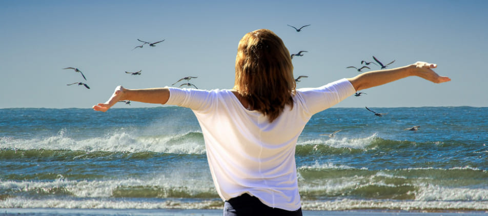 Rückenansicht einer jungen Frau am Strand, die ihre Arme ausreitet und Wellen und Möwen beobachtet. Ganz enspannt nach Schuldenbereingung Privatinsolvenz Schleswig